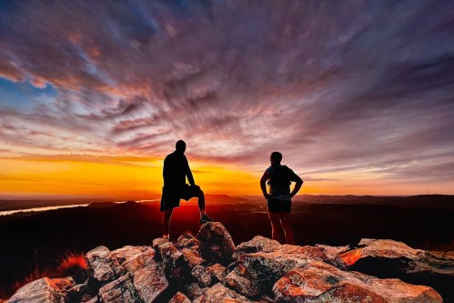 Silhouettes of two people on a rocky outcrop, watching a vibrant sunset over a valley.