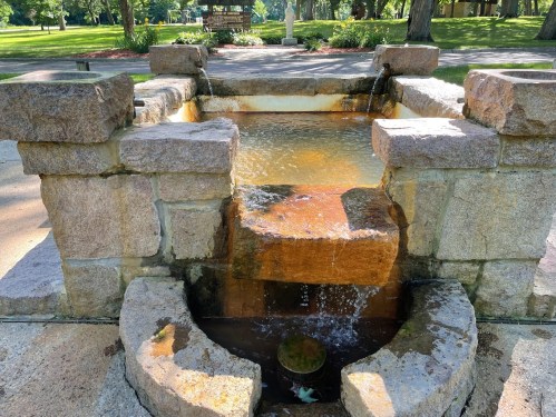 A stone fountain with water flowing over steps, surrounded by greenery in a park setting.