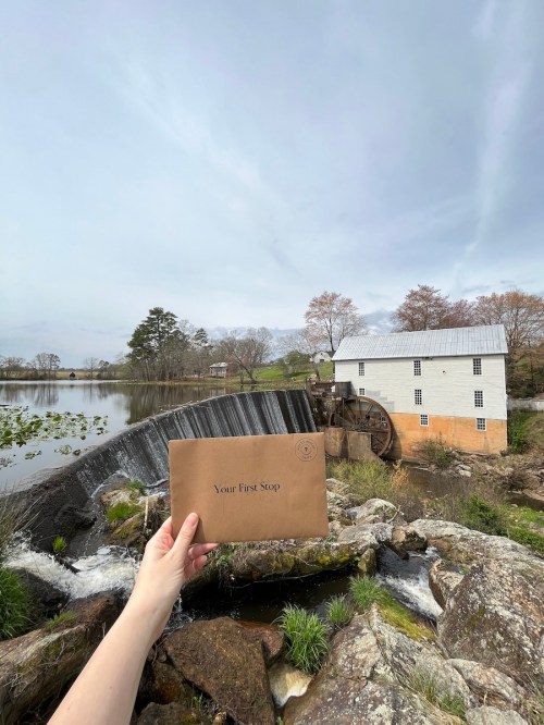A hand holds a brown envelope in front of a scenic watermill and pond, with trees and a cloudy sky in the background.