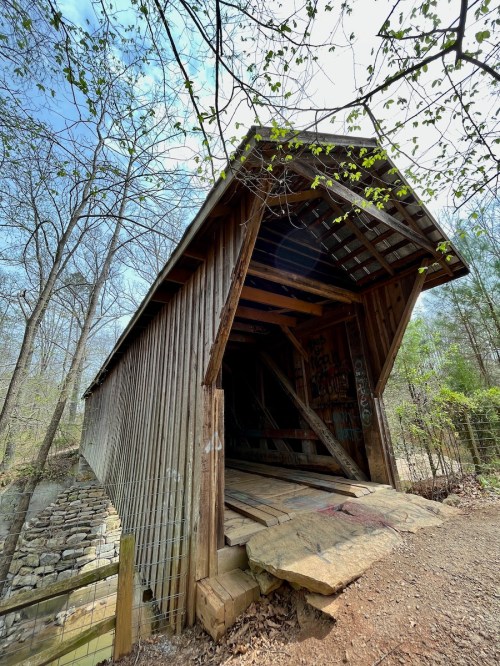 A wooden covered bridge with a sloped roof, surrounded by trees and greenery, leading over a small creek.