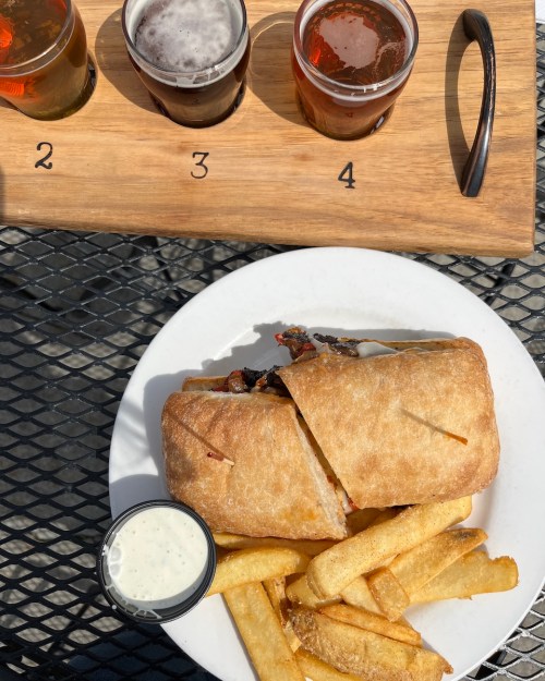 A plate with a sandwich, fries, and a small cup of dipping sauce, alongside a flight of four craft beers.