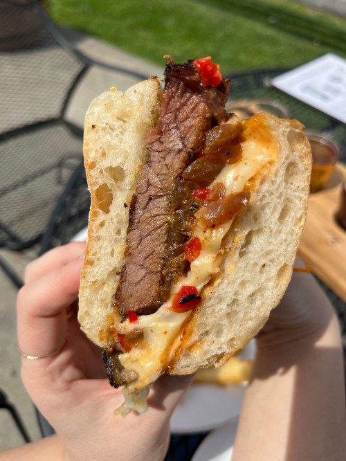 A close-up of a hand holding a sandwich filled with sliced beef, melted cheese, and peppers, with a sunny outdoor setting.