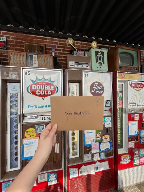 A hand holds an envelope in front of vintage vending machines displaying various drinks and snacks.