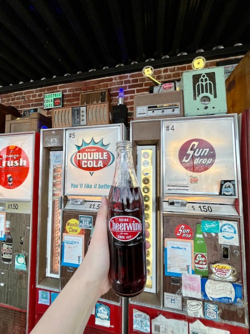 A hand holds a Cheerwine soda bottle in front of vintage vending machines displaying various drinks.