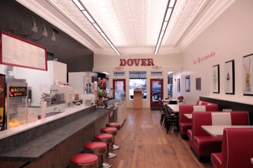 Interior of a diner with red and white decor, a counter, stools, and framed artwork on the walls. "DOVER" sign above.