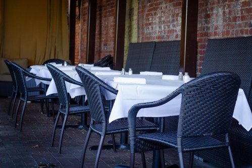 Empty outdoor dining area with wicker chairs and white tablecloths, set against a brick wall.