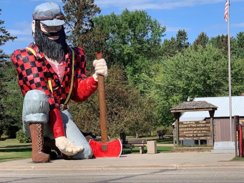 A giant lumberjack statue kneels beside a park sign, surrounded by trees and a clear blue sky.