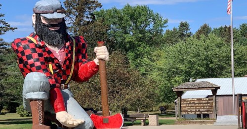 A large statue of a lumberjack in a red and black checkered shirt, holding an axe, in a park setting.