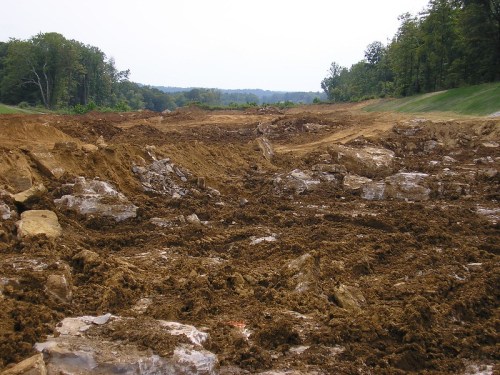 A large, uneven area of disturbed earth and rocks, surrounded by trees and a cloudy sky.