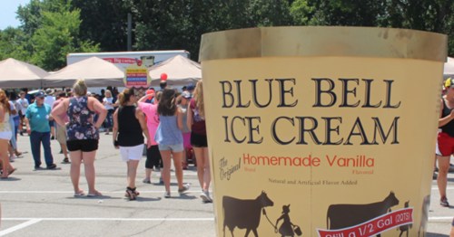 A large Blue Bell Ice Cream container in front of a crowd at an outdoor event, with food trucks in the background.