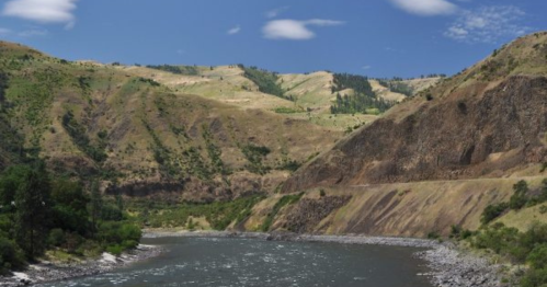 A serene river flows through a mountainous landscape with green hills and rocky cliffs under a clear blue sky.