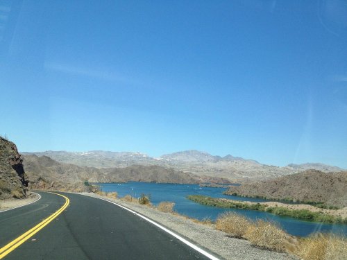 A winding road alongside a blue river, with mountains in the background under a clear blue sky.