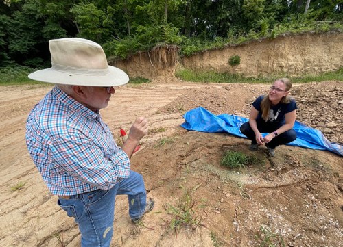 A man in a hat talks to a woman sitting on the ground, surrounded by dirt and greenery, with a blue tarp nearby.
