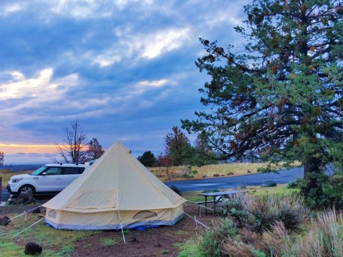 A beige tent set up near a tree, with a car parked nearby and a cloudy sky at sunset in the background.