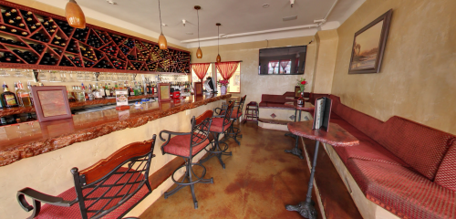 A cozy bar interior featuring a marble counter, red seating, and a wall of wine bottles. Warm lighting creates an inviting atmosphere.