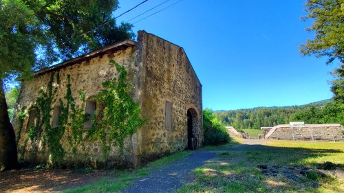 A rustic stone building covered in greenery, set against a clear blue sky and lush green landscape.