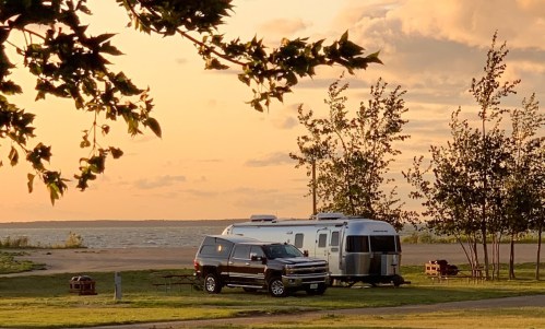 A silver Airstream trailer parked by a lake at sunset, with a dark SUV nearby and trees framing the scene.