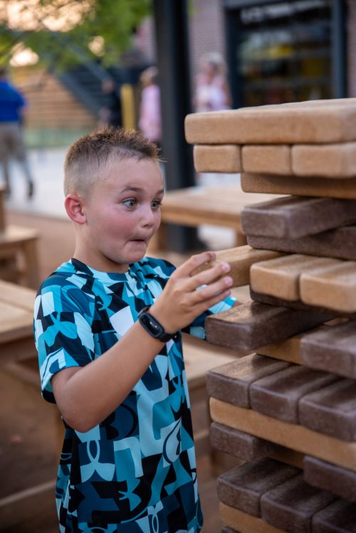 A surprised boy in a patterned shirt reaches for a block in a large outdoor stacking game.