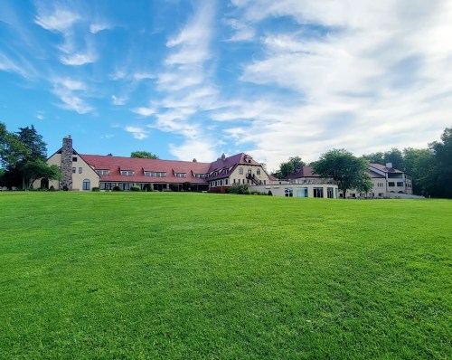 A large, elegant building with a red roof sits on a green lawn under a blue sky with scattered clouds.