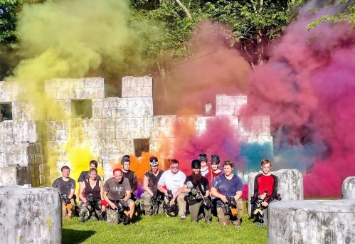 A group of people in tactical gear poses in front of colorful smoke clouds at an outdoor paintball arena.