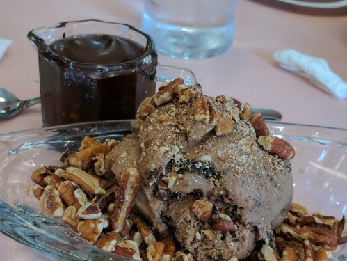 A bowl of chocolate ice cream topped with chopped pecans, alongside a small pitcher of chocolate sauce.