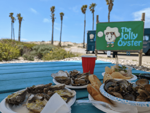 A beachside table with plates of oysters and seafood, a red cup, and a sign for "The Jolly Oyster" in the background.