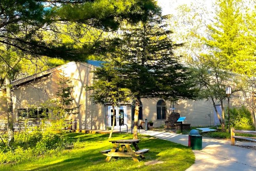 A sunny outdoor view of a building surrounded by trees, with a picnic table and pathway leading to the entrance.
