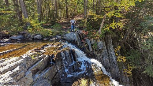 A person stands on rocks by a waterfall in a forested area, surrounded by trees and autumn foliage.