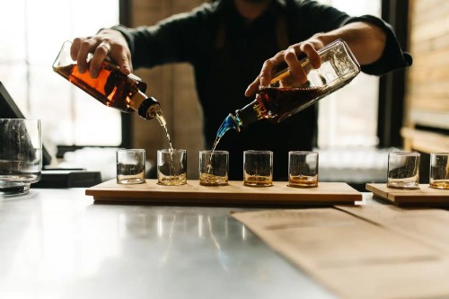 A bartender pours whiskey from two bottles into six glasses on a wooden tray in a stylish bar setting.