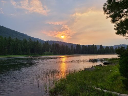 A serene lake at sunset, surrounded by mountains and lush trees, with reflections shimmering on the water's surface.