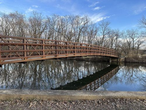 A wooden bridge arches over a calm pond, reflecting trees and a blue sky in the water below.