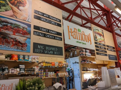 Interior of a vegan deli featuring a menu board and shelves stocked with various food items.