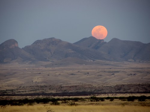 A large orange moon rises over rugged mountains and a vast, dry landscape at dusk.