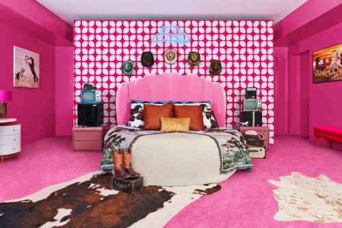 A vibrant pink bedroom featuring a large bed, patterned wall, vintage TVs, and decorative items on a cowhide rug.