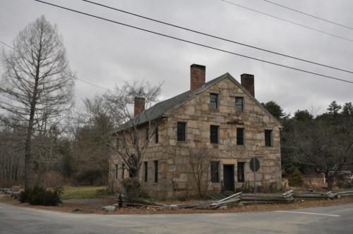 Abandoned stone house at a rural intersection, surrounded by trees and overgrown vegetation under a cloudy sky.