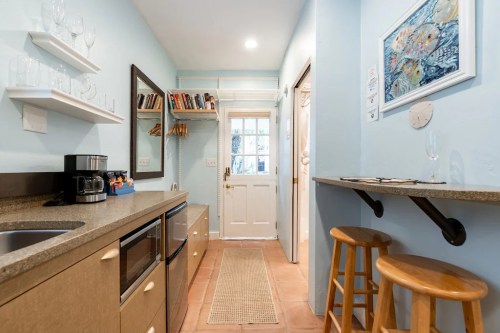 Bright kitchen with light blue walls, a small bar with stools, and a door leading outside. Shelves hold books and glasses.