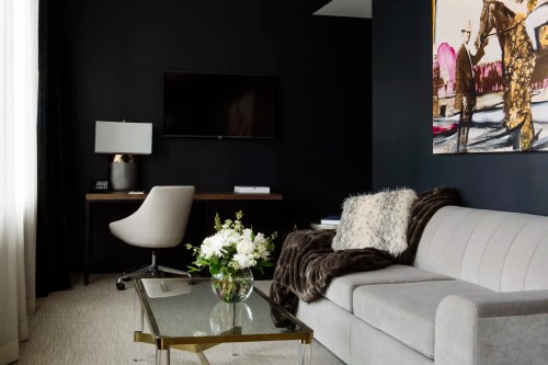 A modern living space featuring a gray sofa, glass coffee table, desk with a chair, and a dark wall with artwork.