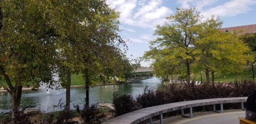 A serene park scene featuring a calm waterway, lush trees, and a cloudy sky. Benches line the pathway.
