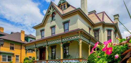 Victorian-style house with colorful trim, a large porch, and blooming flowers in the foreground under a blue sky.