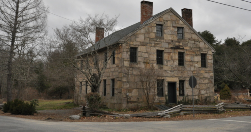 Abandoned stone house with a gray roof, surrounded by trees and a weathered fence on a cloudy day.