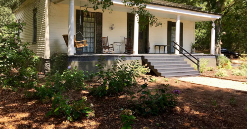 A charming white house with a porch, rocking chairs, and a landscaped garden surrounded by trees.