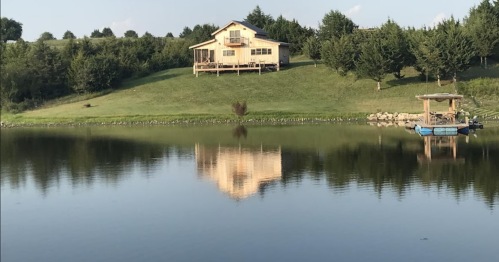 A serene lake reflects a yellow house on a hill, surrounded by trees and a small dock on the water.