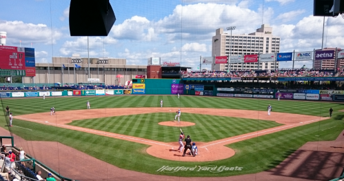 A sunny day at a baseball stadium with players on the field and spectators in the stands. Hartford Yard Goats logo visible.