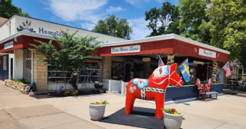 A colorful Dala horse statue stands outside a shop with a red and white facade, surrounded by greenery and decorations.