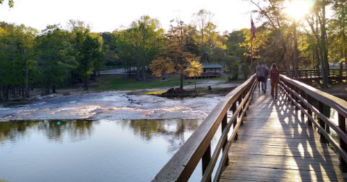 A wooden bridge over a river at sunset, with two people walking and trees lining the banks.