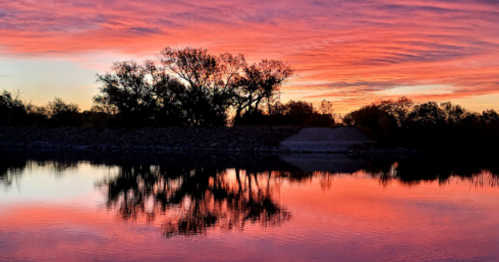 A serene sunset over a calm river, with vibrant pink and purple skies reflecting on the water and silhouetted trees.