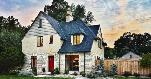 A charming two-story house with a stone facade, dark roof, and red door, surrounded by greenery at sunset.