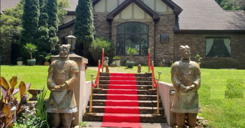 A red carpet leads up stone steps flanked by two large warrior statues in front of a stone house.