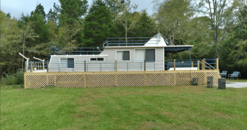 A unique house on stilts with a deck, surrounded by trees and a grassy area.