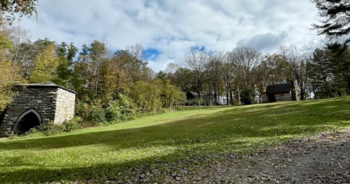 A grassy area with trees, a stone structure, and a small building under a partly cloudy sky.